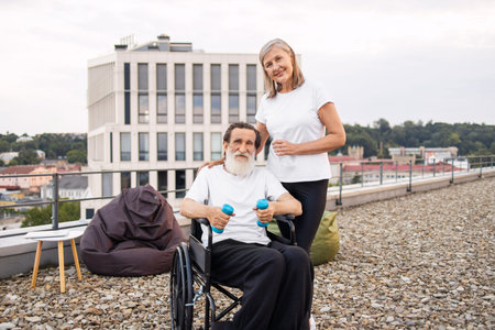 Elderly woman assists wheelchair-bound husband with exercise using dumbbells outdoors, promoting recovery and wellness.の写真素材