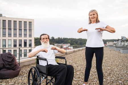 Elderly man with disability in wheelchair and senior woman stretching arms outdoors on rooftop....