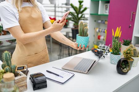 A Caucasian woman is taking a photo of a cactus at a florist shop. There is a cash register, laptop, and other plants on the counter.の写真素材