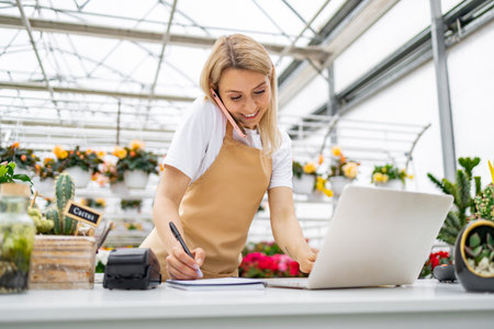 A Caucasian woman working in a florist shop, taking a call and using a laptop while writing.の写真素材