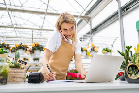 A Caucasian woman, working at a flower shop, is on the phone, taking notes, and using a laptop on a bright day.の写真素材