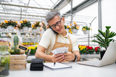 A Caucasian florist on the phone counts cash in his flower shop with a laptop and plants in the background.の写真素材