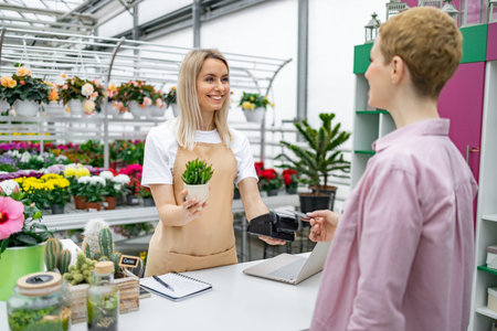 A florist helps a customer pay for a purchase at the checkout, with a smile, holding a small plant...