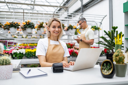A florist is at work, smiling and using a laptop. In the background, a colleague is inspecting a plant, in a sunny greenhouse filled with flowers.の写真素材