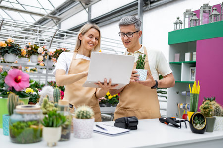 Two florists, a man and a woman, look at a laptop in a plant shop, discussing a small plant in a pot at the table.の写真素材