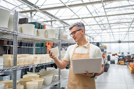 A Caucasian man inspects a flowerpot in a plant shop, holding a laptop for inventory. The shop is organized with shelves full of various pots.の写真素材