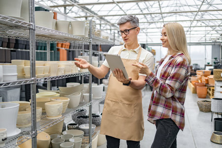 A Caucasian woman discusses flower pots with a shop assistant at a plant store. Theyre examining options on a tablet device near shelves.の写真素材