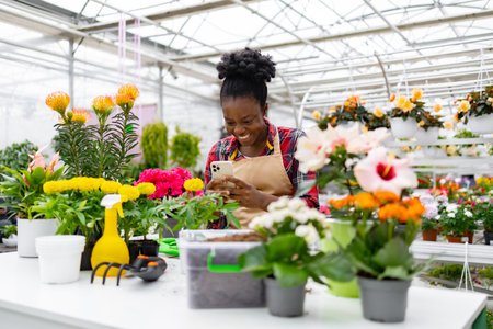 A smiling Black woman in a greenhouse looks at her phone surrounded by colorful flowers.の写真素材