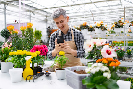 A smiling man in an apron uses his phone while standing in a vibrant greenhouse filled with a variety of colorful flowers and plants.の写真素材
