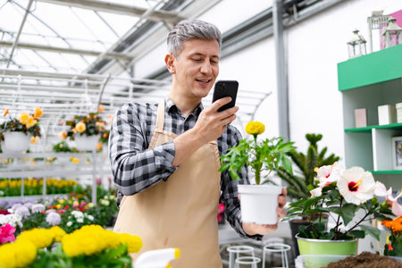 A man in a greenhouse takes a photo of a potted plant, surrounded by various flowers, using his phone.の写真素材