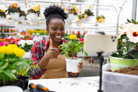A cheerful Black woman in a greenhouse holds a plant, giving a thumbs-up while recording a video on her phone.の写真素材