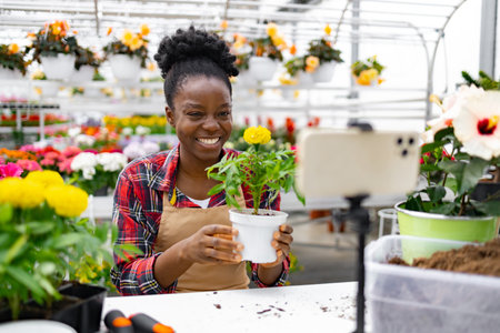 A smiling Black woman holds a potted plant, recording a video in a vibrant greenhouse filled with colorful flowers.の写真素材