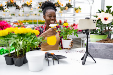 A woman in a greenhouse demonstrates plant care, holding a potted flower and a spray bottle while recording with her phone.の写真素材