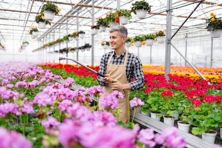A man waters flowers in a vibrant greenhouse filled with colorful blooms and hanging plants.の写真素材