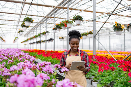 An African American woman smiles while using a tablet in a colorful flower greenhouse, surrounded by blooming plants.の写真素材