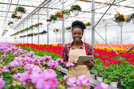 A smiling African woman in a greenhouse with a tablet, surrounded by vibrant flowers and plants, and other potted plants.の写真素材