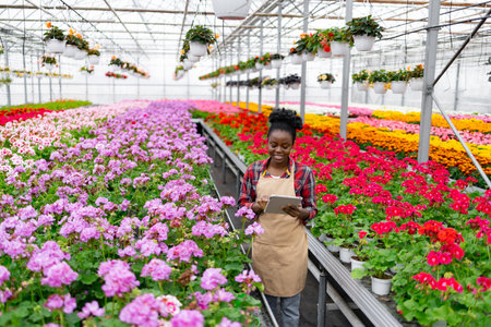 A smiling African woman in a greenhouse manages inventory on a tablet, surrounded by vibrant flowers and hanging plants.の写真素材