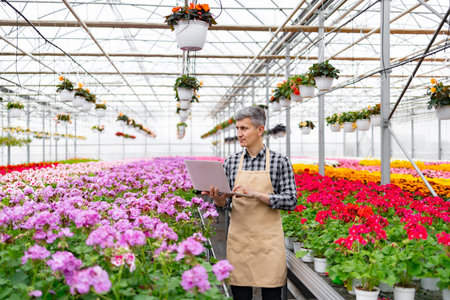 A gardener in a greenhouse, using a laptop to manage vibrant flowers and plants in pots and hanging baskets.の写真素材