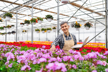 A smiling greenhouse worker examines flowers with a laptop, surrounded by vibrant blooms in a colorful floral arrangement.の写真素材