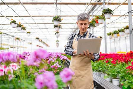 A man in a greenhouse, working on a laptop amidst vibrant flowers, managing plants.の写真素材