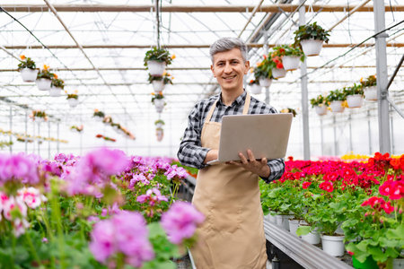 A smiling florist stands in a greenhouse, using a laptop to manage the plants.の写真素材