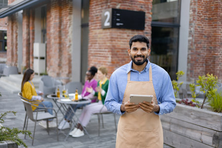 Smiling Hindu male waiter holding a tablet outdoors, service in a restaurant.の写真素材