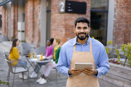 A smiling Hindu male waiter in an apron holds a tablet, standing outside a restaurant.の写真素材