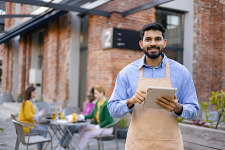 A smiling Hindu male waiter holds a tablet while looking at the camera, apron on, outdoors.の写真素材