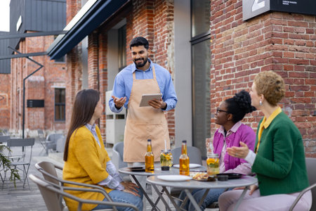 A waiter takes orders on a tablet from a group of women seated outdoors at a cafe.の写真素材