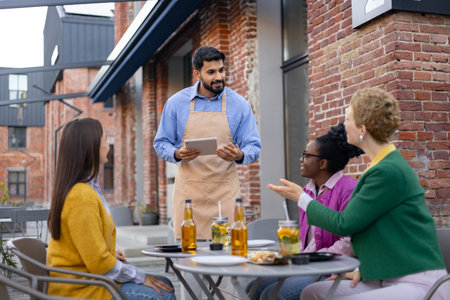 A waiter takes an order on a tablet at an outdoor cafe. Three women sit at the table, socializing.の写真素材