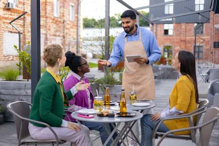 A waiter takes an order from three women seated at an outdoor restaurant table. They are enjoying drinks and conversation.の写真素材