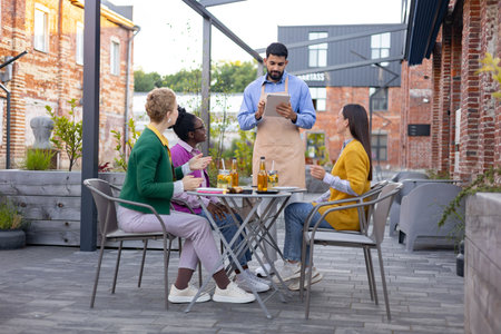 A waiter takes an order from a group of friends sitting at an outdoor cafe table, enjoying drinks and conversation.の写真素材