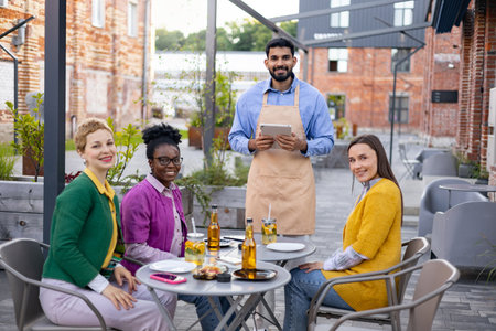 A diverse group of friends enjoys a meal at an outdoor cafe, served by a friendly waiter.の写真素材