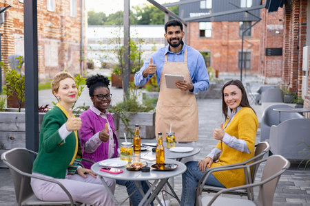 Happy group of friends and a waiter giving thumbs up in a restaurant outdoor setting.の写真素材