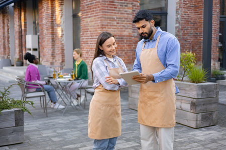A diverse pair of restaurant staff using a tablet while discussing something outside a brick building.の写真素材