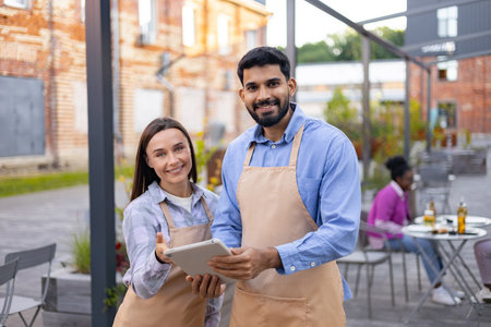 Smiling cafe workers holding a tablet and standing outdoors, with customers in the background.の写真素材