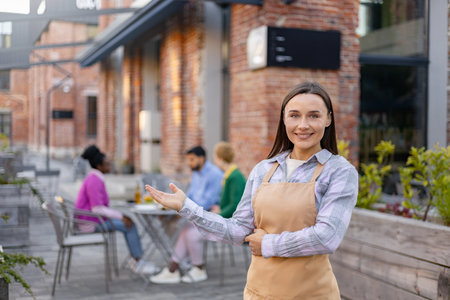 A smiling waitress in an apron welcomes customers to an outdoor restaurant. Other patrons are seated at tables in the background.の写真素材