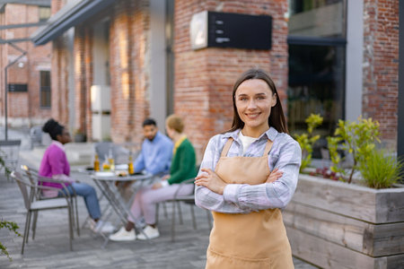 Smiling waitress standing arms crossed at a restaurant patio, with customers at a table in the background.の写真素材