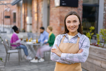 A smiling waitress in an apron poses in front of her outdoor restaurant, customers sit at tables in the background.の写真素材