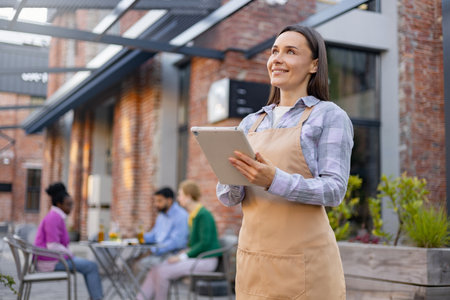 A waitress holding a tablet, smiles while looking up, with restaurant patrons in the background.の写真素材