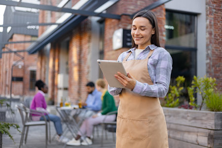 A smiling waitress takes an order at an outdoor restaurant with a tablet, overseeing customers.の写真素材