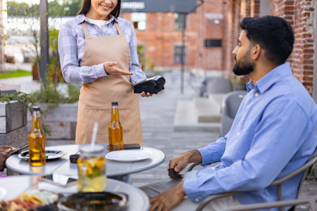 A female waiter offers a payment terminal to a customer at an outdoor restaurant table.の写真素材