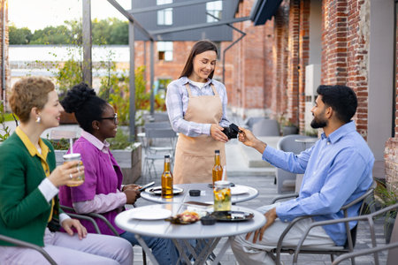 A female waiter assists customers with a portable terminal for payment at an outdoor cafe.の写真素材