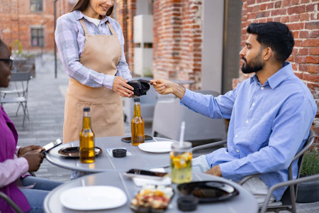 A female waiter at an outdoor cafe assists a customer with payment using a portable terminal.の写真素材