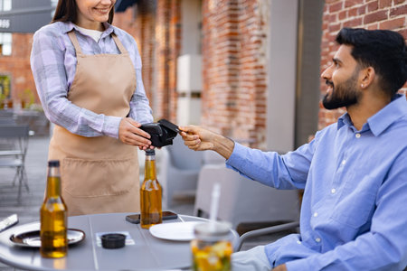A smiling female waiter presents a payment terminal to a male customer seated at an outdoor cafe table.の写真素材