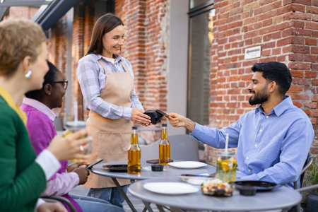 A female waiter is at a table and is asking a customer to pay the bill using a portable payment terminal. Other customers are present.の写真素材