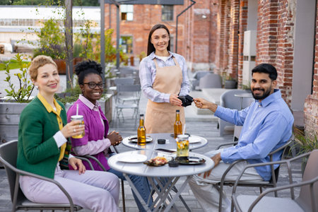 A female waiter in an apron serves customers at an outdoor restaurant, accepting payment via a portable terminal.の写真素材