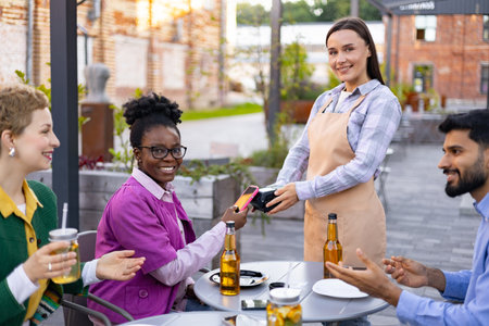 A female waiter in an apron uses a portable terminal to process a payment at an outdoor restaurant, customers are ready to pay.の写真素材