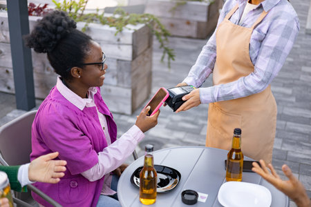 A female waiter using a portable terminal, assisting a customer with a payment in the outdoor cafe.の写真素材
