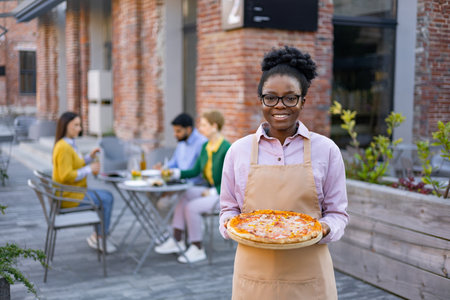 Smiling African waitress holding a pizza, serving customers in an outdoor restaurant setting.の写真素材